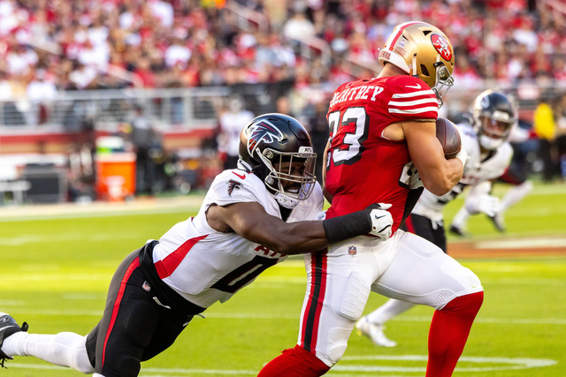 A football player in a red San Francisco 49ers uniform clutches the ball while a player in a white Atlanta Falcons uniform tackles him from behind.